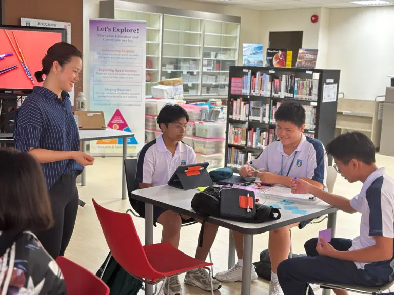 hod-briefing-design-thinking-workshop-singapore - C-Academy Teacher standing and smiling as students work on tablets and notes at a round table in a library.