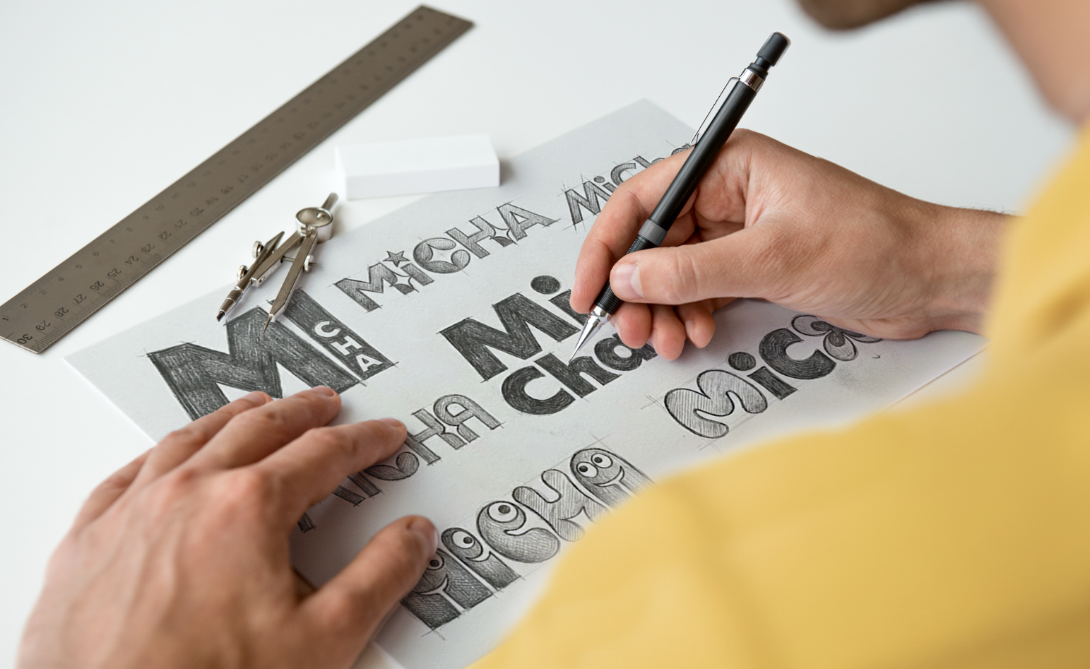Hands sketching a decorative wordmark on white paper, with a ruler, compass, and eraser nearby.