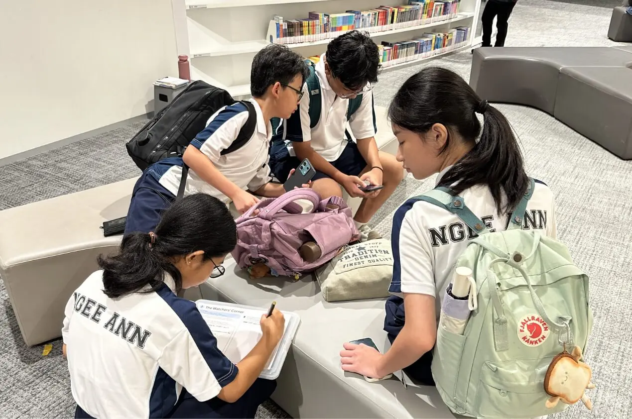 A group of students in matching uniforms sit on a curved lounge in a library, studying with notebooks and backpacks nearby.