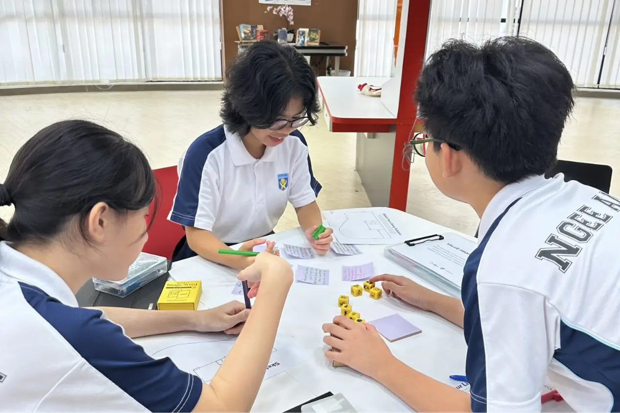 Three students in white-and-blue uniforms collaborate at a table in a bright classroom, using sticky notes, paper, and yellow dice for a project activity.