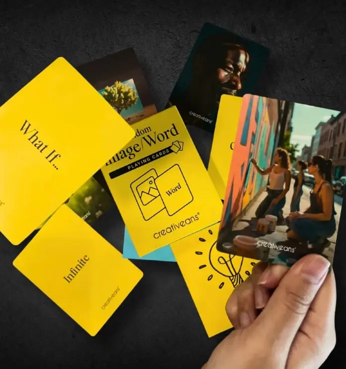 Close-up of a hand holding a word-themed playing card; bright yellow cards with phrases like 'What If...' and 'Infinite' scattered on a dark surface, suggesting a card game.