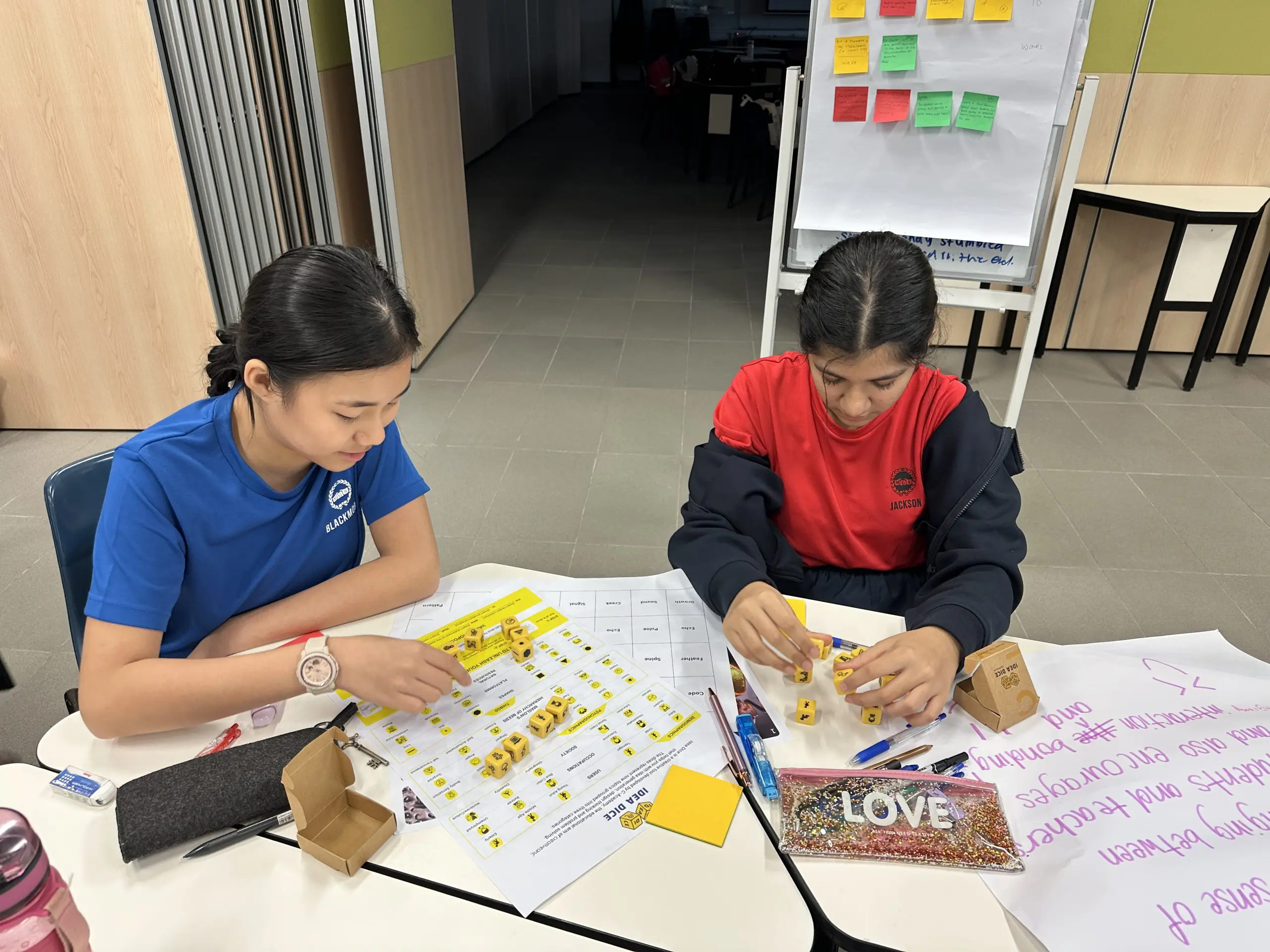 Two children sit at a table working with yellow dice and worksheets in a classroom setting.