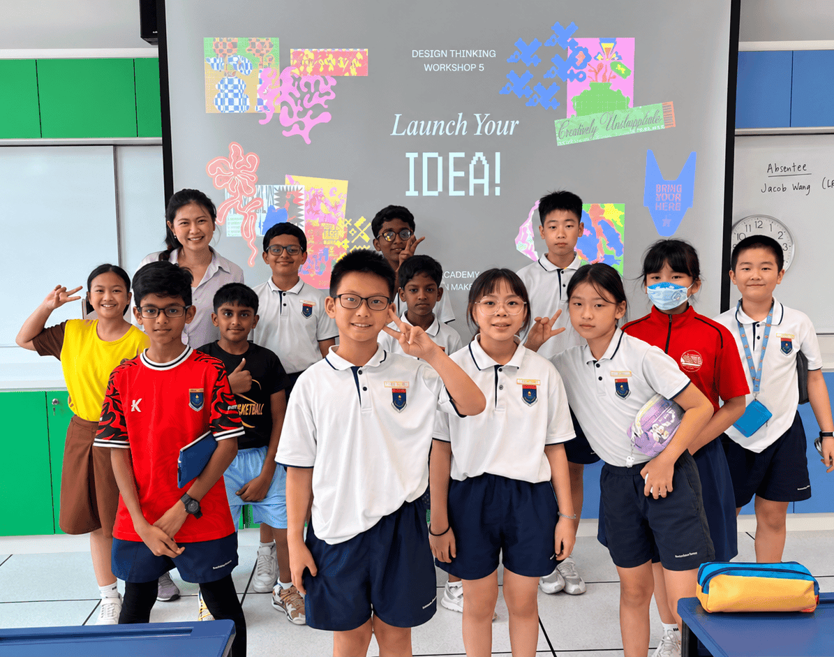 A teacher and a diverse group of students pose for a photo in a classroom with a bright slide that says 'Launch Your IDEA!' in the background.